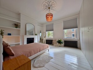 A bedroom with a bed, a fireplace, a mirror, a radiator, and a window with blinds. - Chic Property With Backyard Near Emirates Stadium (London)