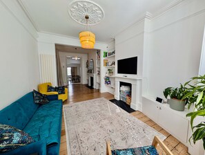 A living room with a blue sofa, a yellow armchair, a chandelier, a fireplace, and a television. - Chic Property With Backyard Near Emirates Stadium (London)