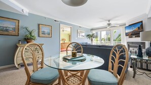 A dining area with a glass table surrounded by wicker chairs, a ceiling fan, and framed artwork on the walls.