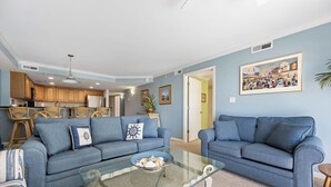 A living room with two sofas, a glass coffee table, and a kitchen in the background.