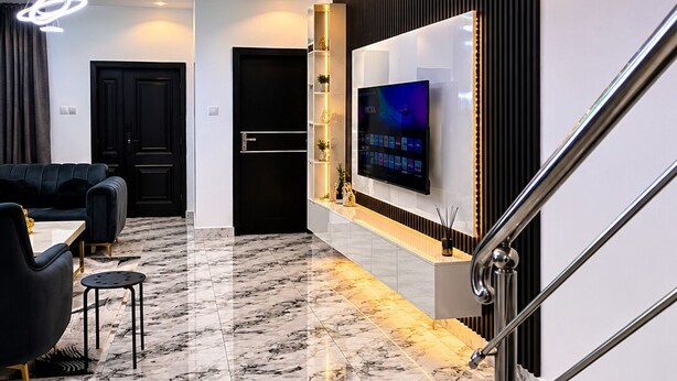 A modern living room with a marble floor, a TV mounted on the wall, and a staircase with metal railings. - Ileero Homes: The Manor | Executive 4-Bedroom (Lagos)