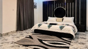 A modern bedroom with a bed, a circular light fixture, a wall-mounted air conditioner, and a patterned rug. - Ileero Homes: The Manor | Executive 4-Bedroom (Lagos)