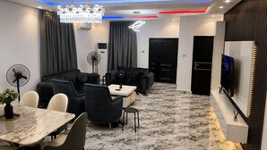 A modern living room with a marble floor, black sofas, a dining table with chairs, a TV, and a chandelier. - Ileero Homes: The Manor | Executive 4-Bedroom (Lagos)
