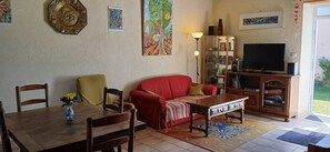 Un salon avec un canapé rouge, une table basse en bois et un téléviseur. On y trouve des fauteuils, une petite table avec un vase et une bibliothÚque.