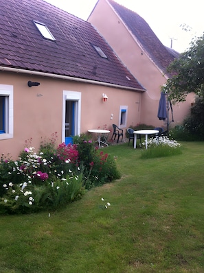 Une maison avec un toit en pente, une petite fenĂȘtre, une porte et un jardin avec des fleurs et de lâherbe.