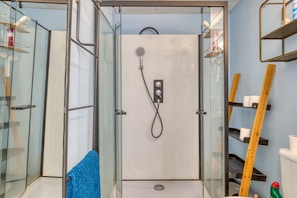 Un espace douche fermé par des parois de verre, doté d’un pommeau de douche à main, d’une serviette bleue et d’une échelle en bois appuyée contre le mur.