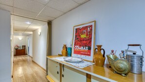 Un couloir avec un plancher en bois, un mur blanc et une affiche. On peut voir des objets décoratifs sur une table, notamment des vases et un récipient en métal.