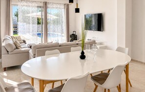Ein modernes Wohnzimmer mit einem weißen Sofa, einem Esstisch mit Stühlen, einem an der Wand befestigten Fernseher und Blick ins Freie.