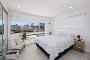 A bedroom with a large bed, a nightstand, a bedside table, a window with a view of buildings, and a small plant. - affordable modern getaway with parks/cafe nearby (WOODPARK)
