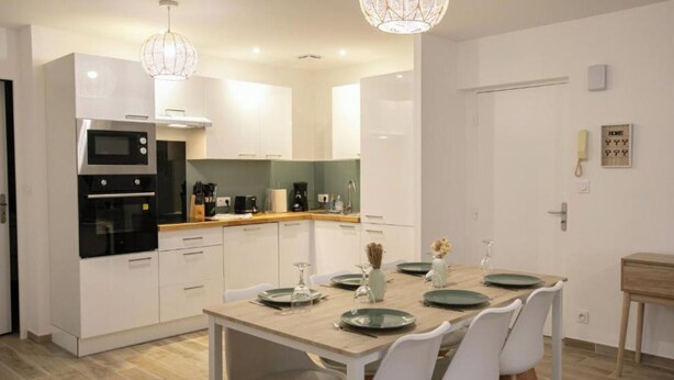 A modern kitchen with white cabinets, a built-in oven, and a dining area with a wooden table and chairs. - Elegant 2-bedroom apartment-Near downtown (Périgueux)