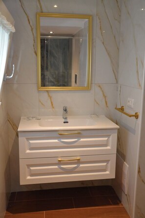 Un bagno con parete in marmo, un mobile vanitario bianco con maniglie dorate, uno specchio con cornice dorata e un pavimento piastrellato scuro.