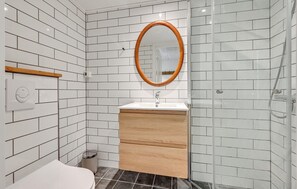Une salle de bain avec des carreaux de céramique blancs rectangulaires, un meuble-lavabo en bois, un miroir rond, une douche et des toilettes.