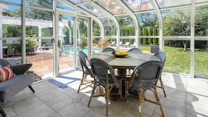 Uma conservatória com teto de vidro, uma mesa de jantar redonda, cadeiras de vime e vista para um jardim e piscina.