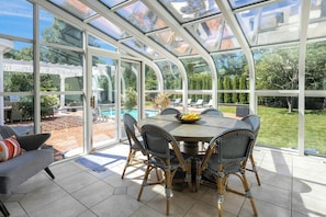Une véranda dotée d’un toit en verre, une table à manger ronde, des chaises en osier et une vue sur un jardin et une piscine.