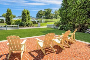 Eine Terrasse mit Adirondack-Stühlen und einem Ziegelboden, mit Blick auf einen See und viel Grün.