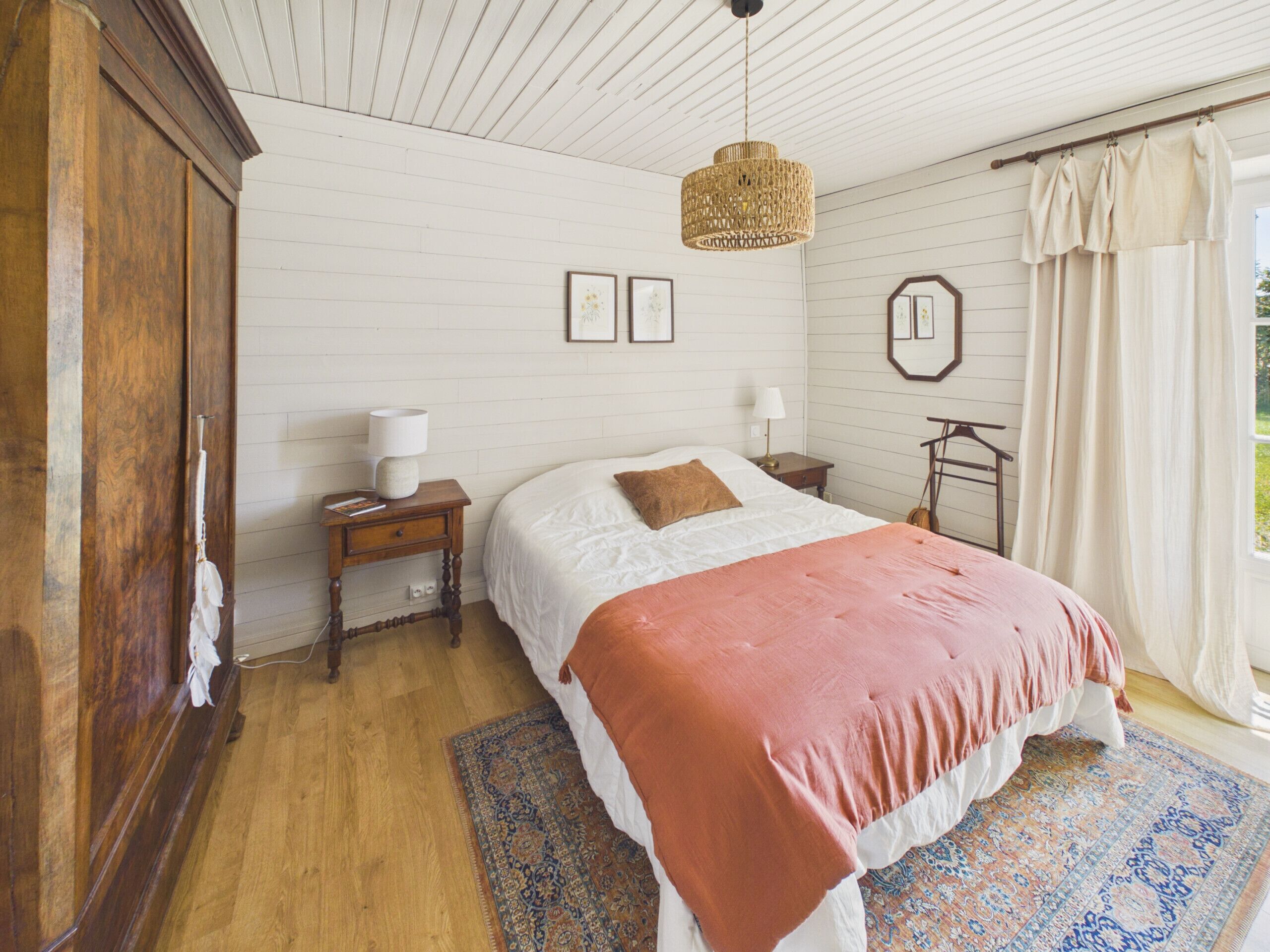 Une chambre avec une armoire en bois, un lit recouvert d’une couvre-lit blanche et orange, un luminaire suspendu en rotin, une table de chevet avec une lampe et un tapis à motifs.