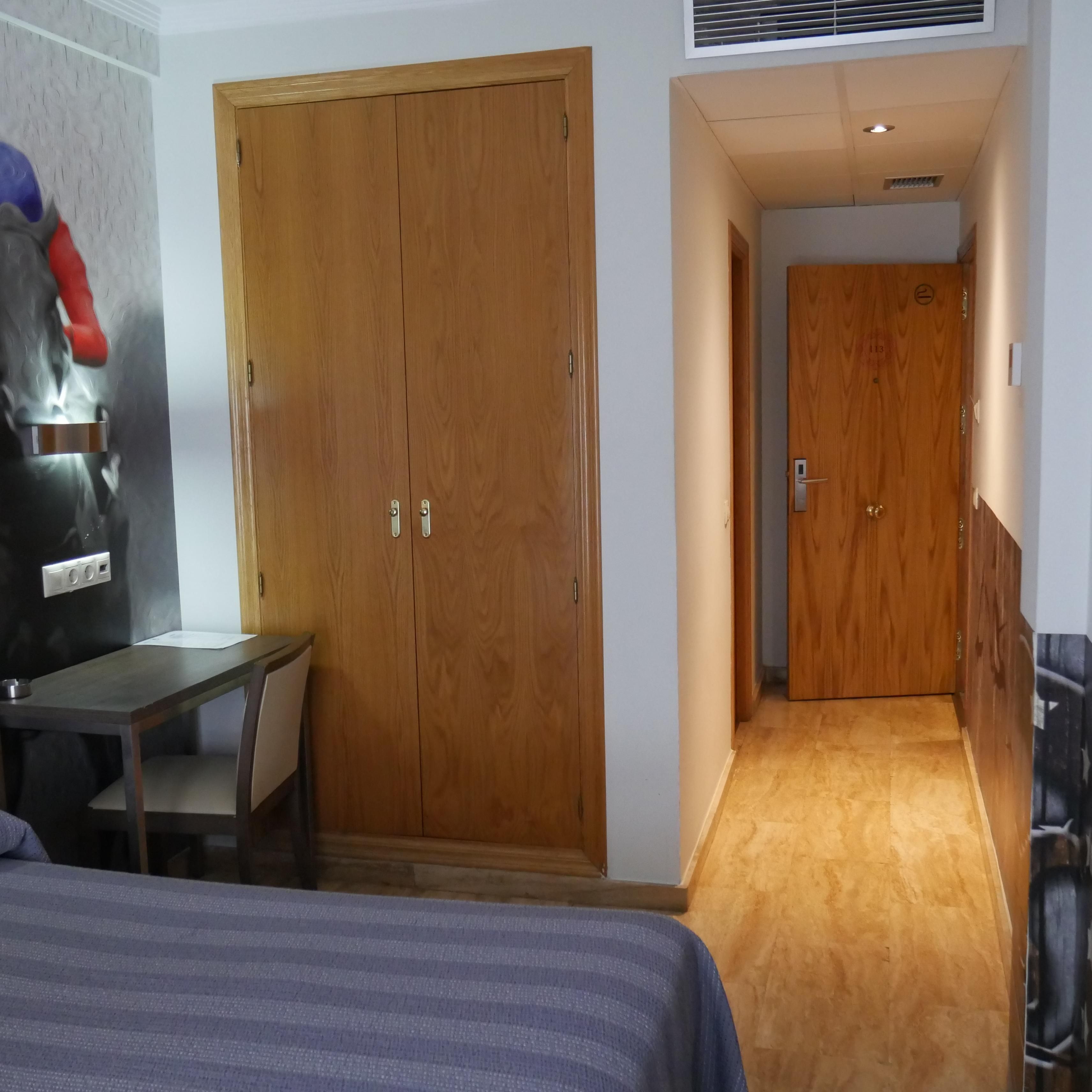 Ett hotellrum med en garderob i trä, ett skrivbord, en säng och en korridor som leder till ett annat rum.
