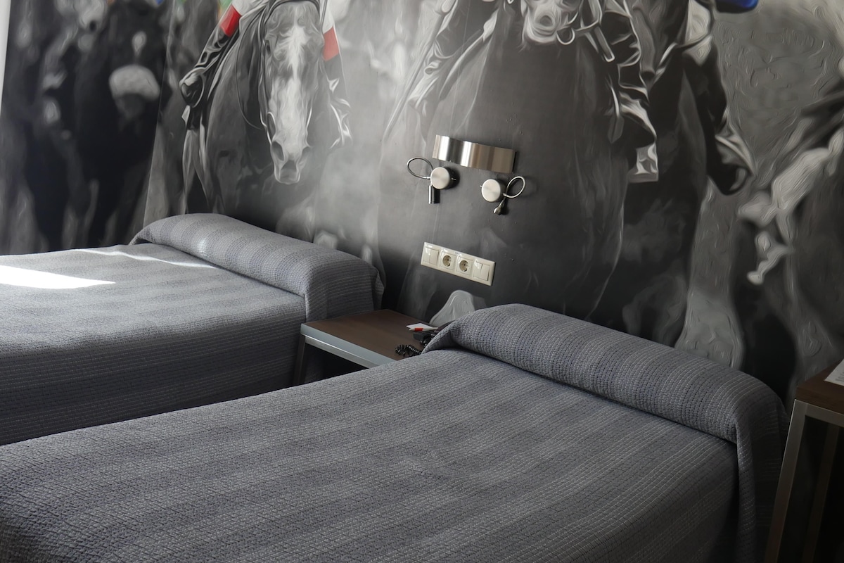 Quarto com um grande mural de jóqueis a cavalo, um sofá cinzento e uma pequena mesa.