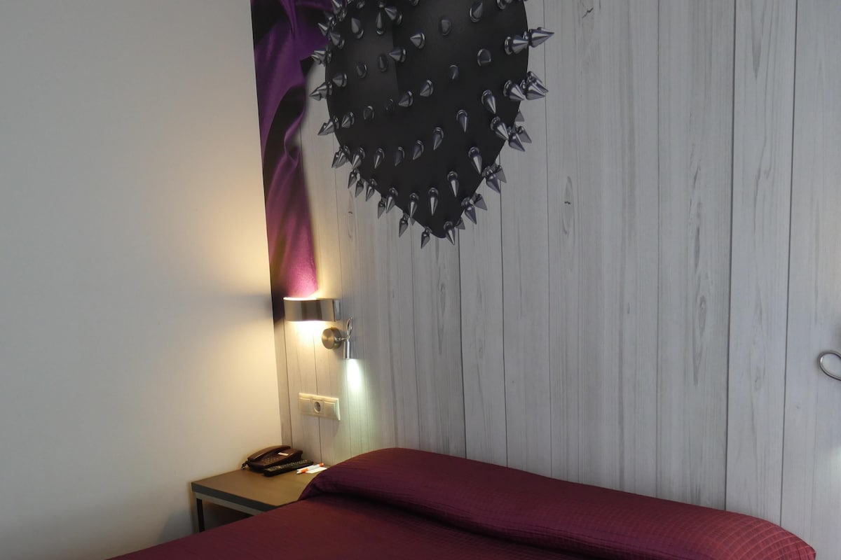 Quarto com cama, mesa de cabeceira, abajur e um enfeite na parede.