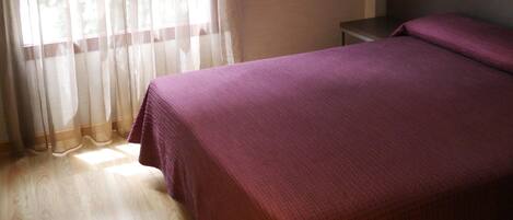 A bedroom with a bed, a nightstand, a wall-mounted lamp, and sheer curtains.