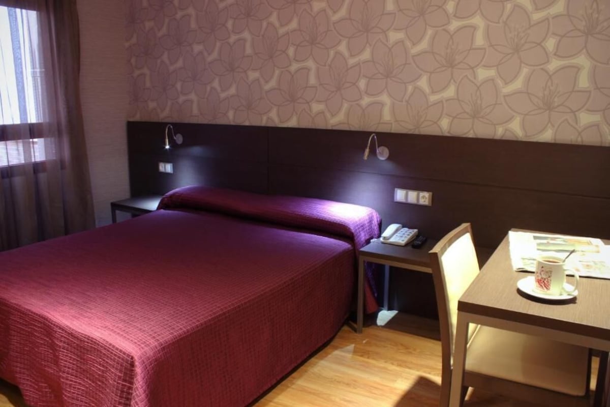 Una camera d'albergo con un letto, un comodino, una sedia e un tavolino con una tazza e dei documenti.