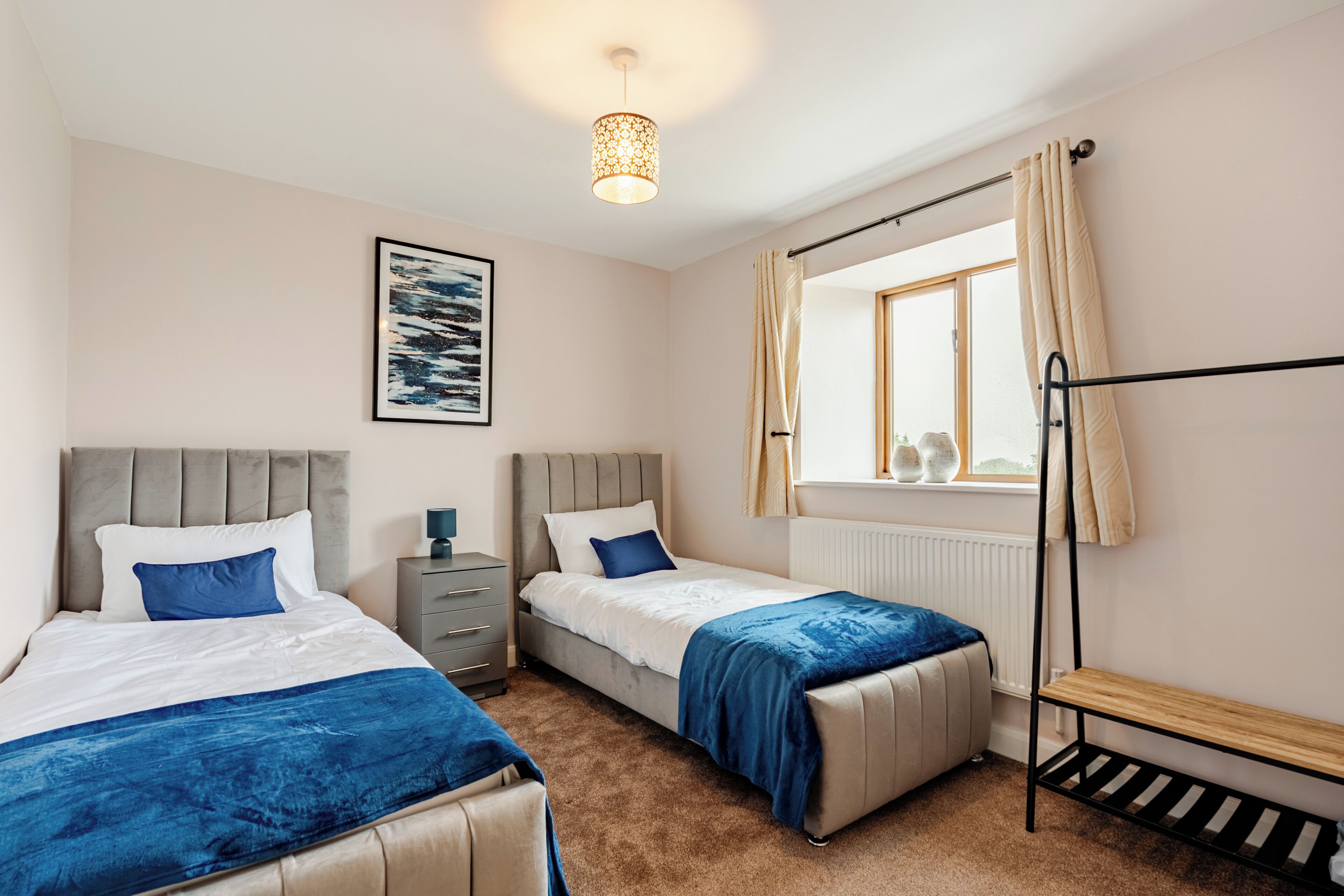 A bedroom with two beds, a bedside table, a lamp, a window with curtains, and a radiator.