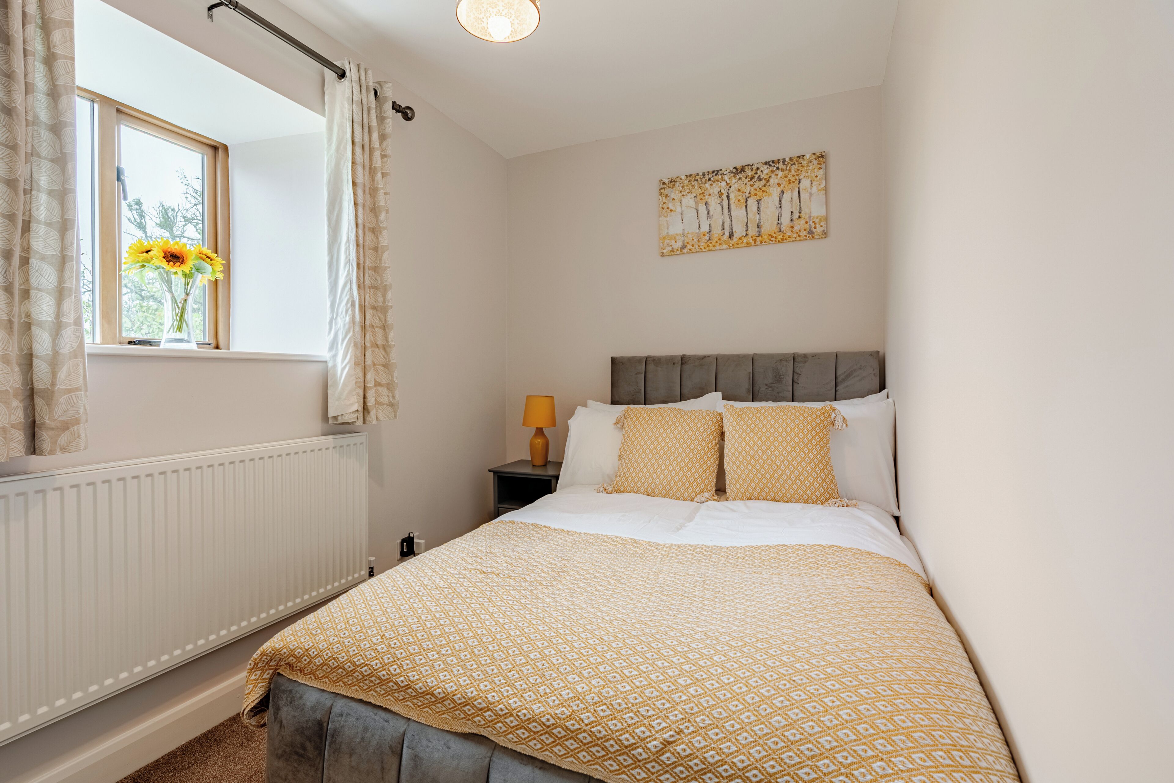 A bedroom with a bed, a window with curtains, a bedside table with a lamp, and a wall-mounted painting.