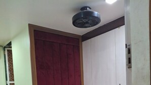 A single bed with a white mattress, two pillows with colorful geometric patterns, a patterned bedspread, a ceiling fan, and a wooden headboard. - Venus Guest House  (Mumbai)
