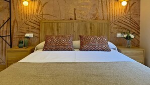 A bedroom with a large bed, two patterned pillows, a wooden headboard, a nightstand with a vase of flowers, and a wall with a leaf pattern. - Casa Las Mironas - III - Ole Solutions (Puerto de Santa María)