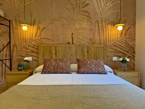 Ein Schlafzimmer mit einem großen Bett, zwei gemusterten Kissen, einem Holzkopfteil, einem Nachttisch mit einer Blumenvase und einer Wand mit einem Blattmuster.