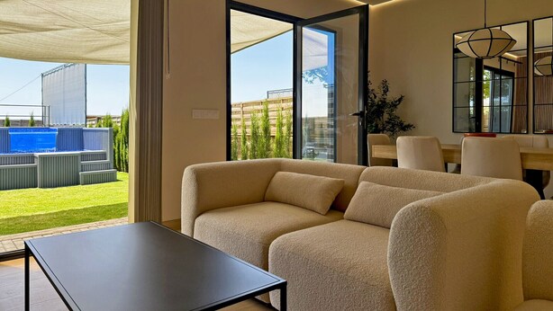 A modern living room with a sofa, a table, and a view of a pool and greenery outside. - Casa Las Mironas - III - Ole Solutions (Puerto de Santa María)