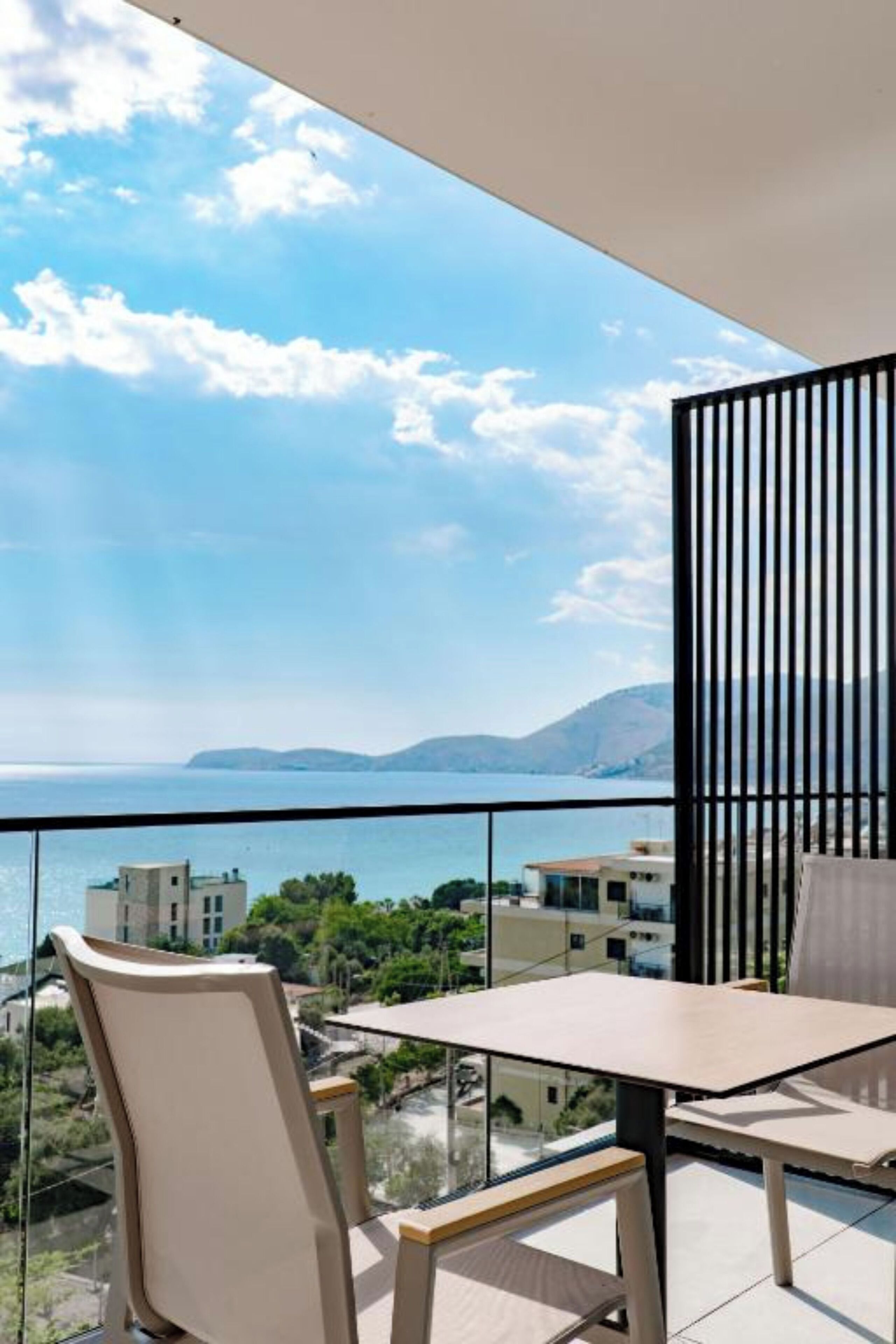 A balcony with a table and chairs, offering a view of the sea and distant buildings.
