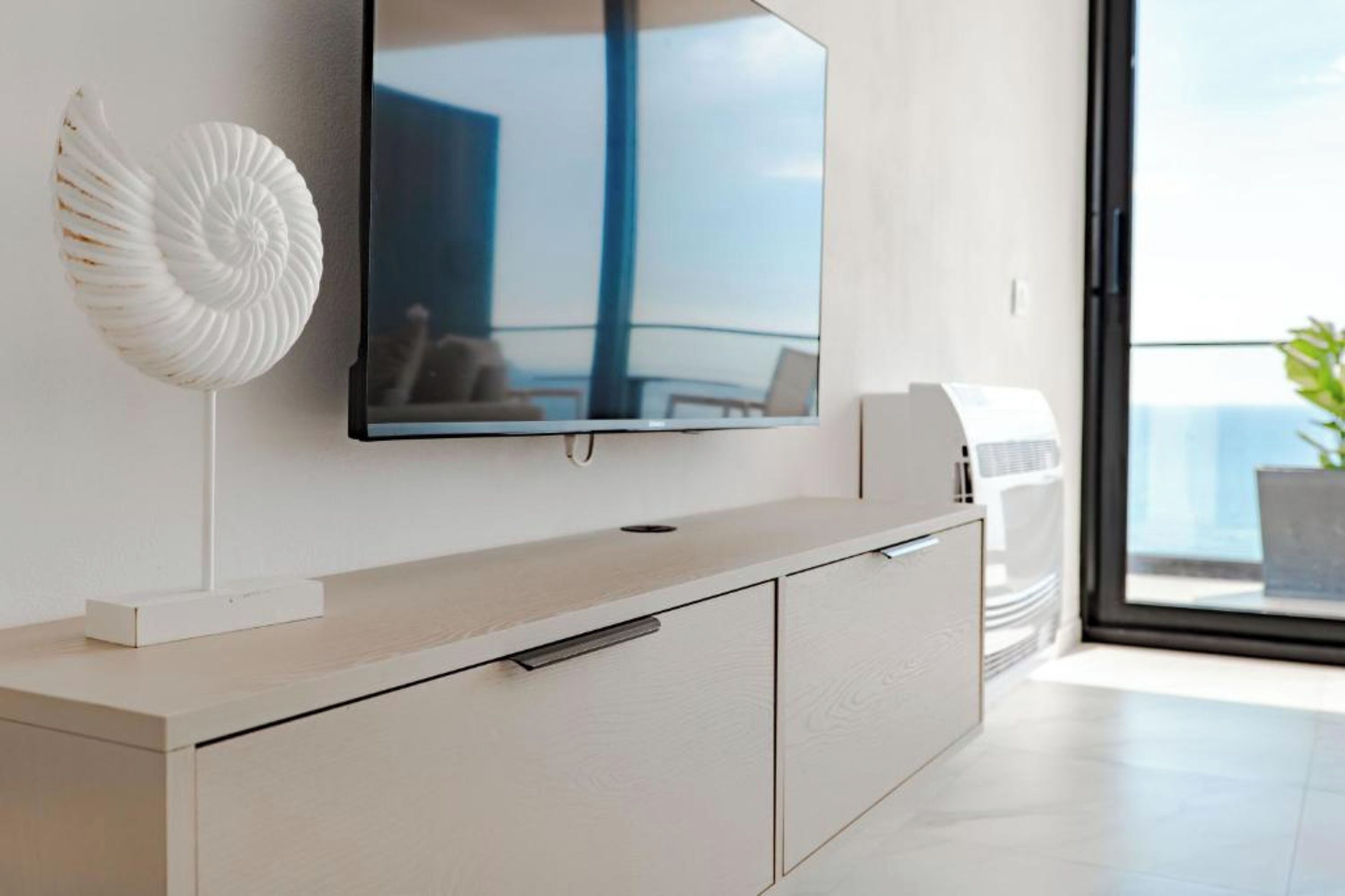 A modern living room with a flat-screen TV mounted on a white cabinet, a decorative shell on the wall, and a view of the outdoors through a large window.
