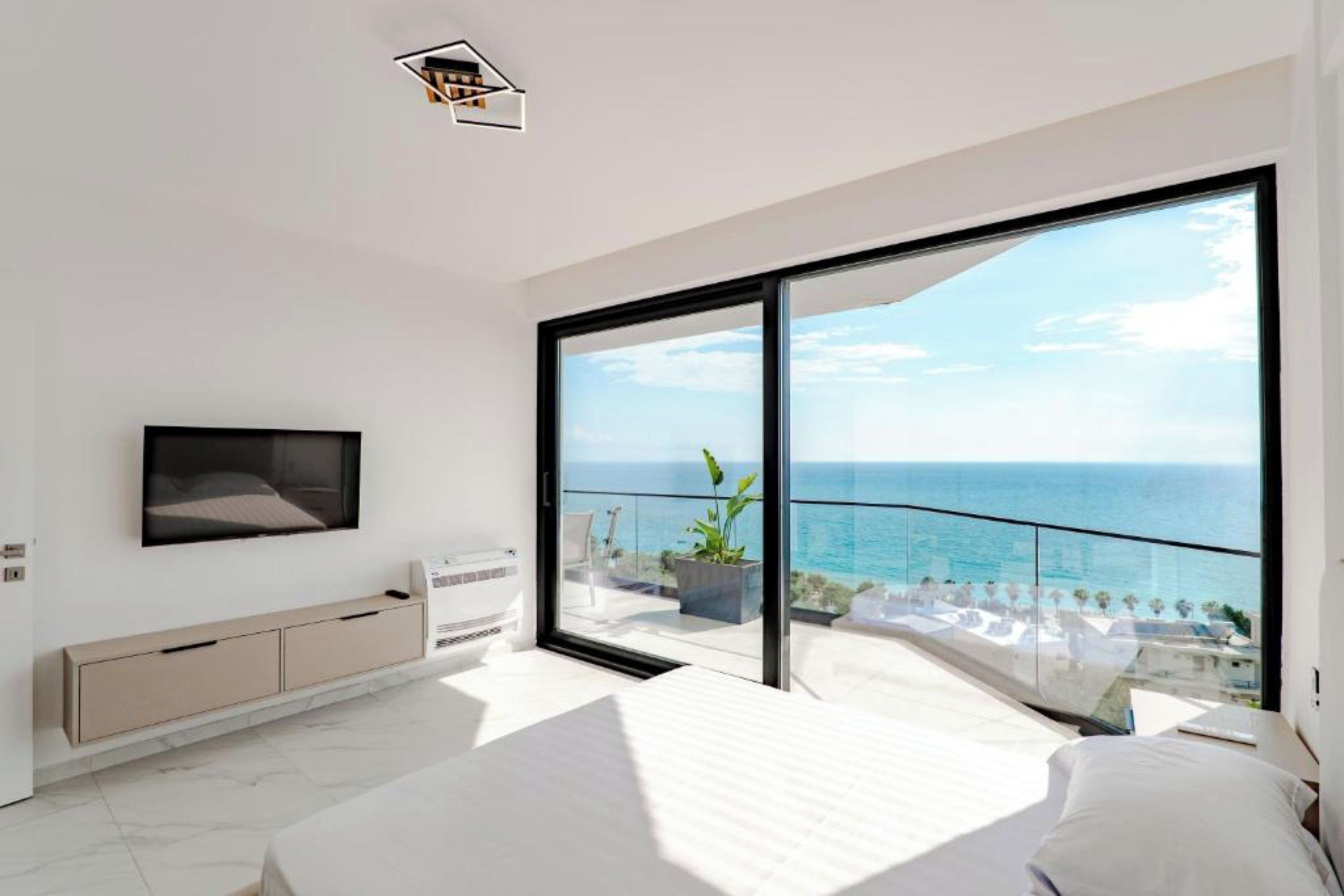 Una camera da letto moderna con un letto grande, una TV fissata alla parete e una porta scorrevole in vetro che dà su un balcone con vista sul mare.