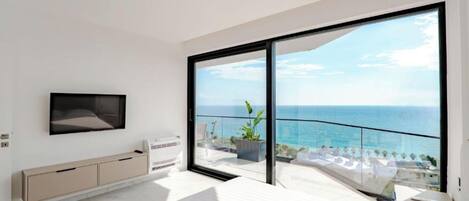 Una camera da letto moderna con un letto grande, una TV fissata alla parete e una porta scorrevole in vetro che dà su un balcone con vista sul mare.