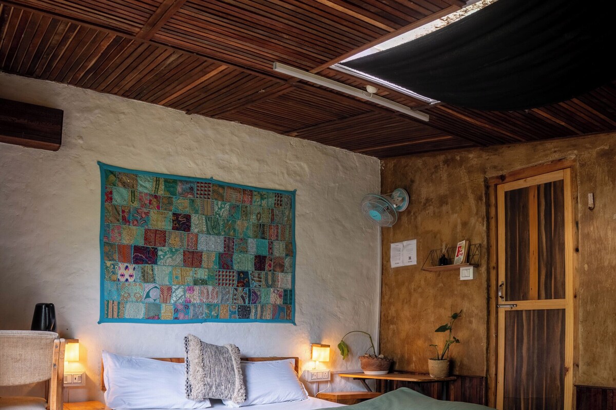 Una habitación acogedora con techo de madera, un tapiz acolchado en la pared, una cama con cubrecama verde, una mesita de noche con lámpara y una puerta de madera.
