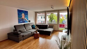 En moderne stue med en grå sofa, et lille rundt bord med en vase, og et stort vindue med udsigt over et boligområde.