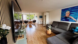 En moderne stue med en grå sofa, et spiseområde med bord og stole, og et stort vindue med udsigt til grøn vegetation.