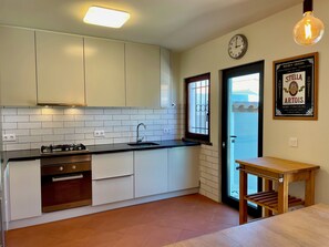 Una cocina moderna con armarios blancos, encimera negra, horno de acero inoxidable, ventana con barrotes, mesa de madera y un reloj en la pared.