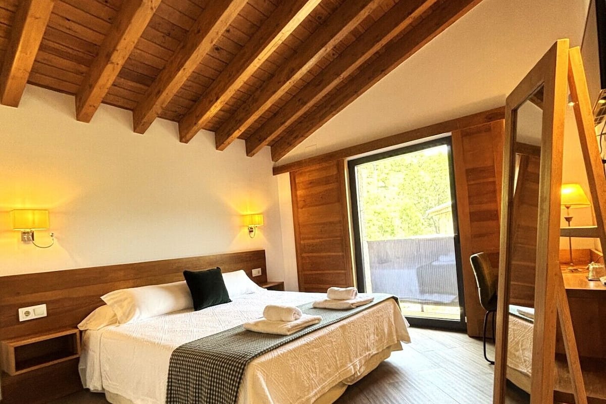 Un dormitorio con techo de madera, una cama con ropa blanca, un ventanal grande y un espejo.