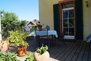 Une terrasse en bois avec une table et des chaises, des plantes en pot et un bâtiment doté d’une fenêtre et de volets.