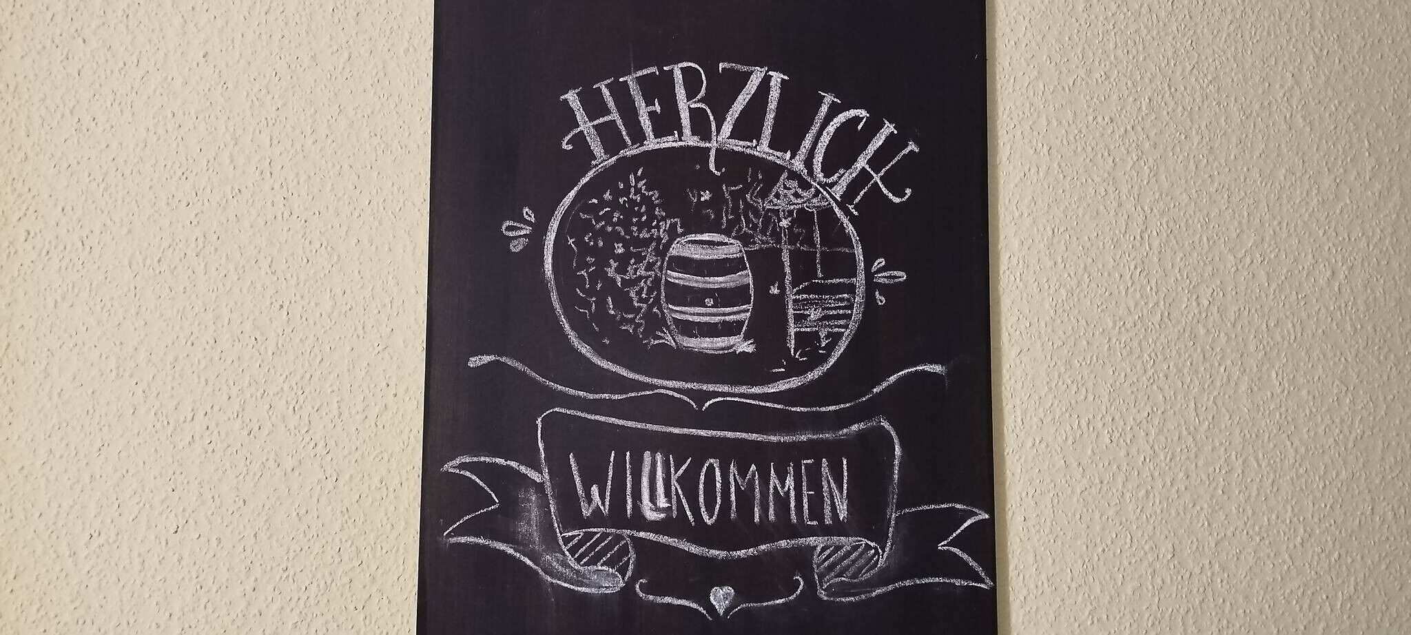A black banner with white chalk drawings of a barrel, a tree, and the words 'HERZLICH WELCOME' on a textured wall.