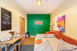 A room with a green accent wall, a bed with floral bedding, a dining area with a table set for two, and a painting on the wall.