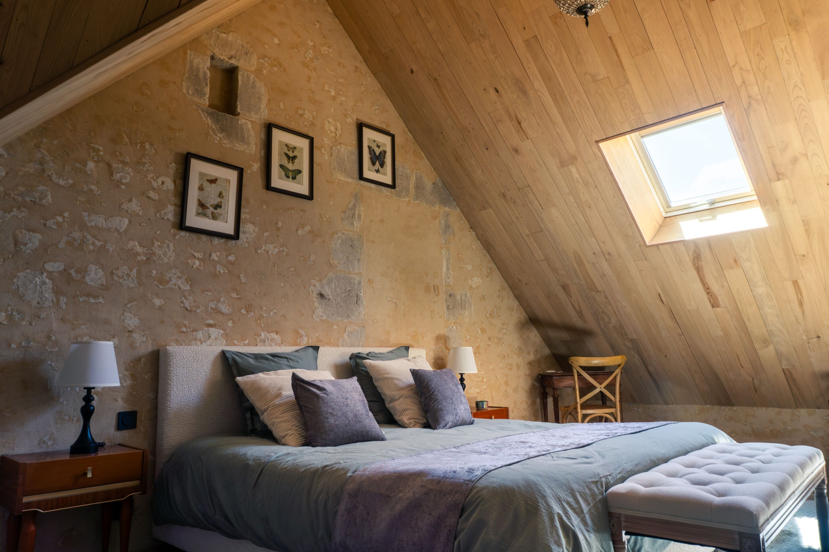 Un dormitorio con techo inclinado, una cama con muchas almohadas, una mesita de noche con una lámpara y una ventana con vistas.