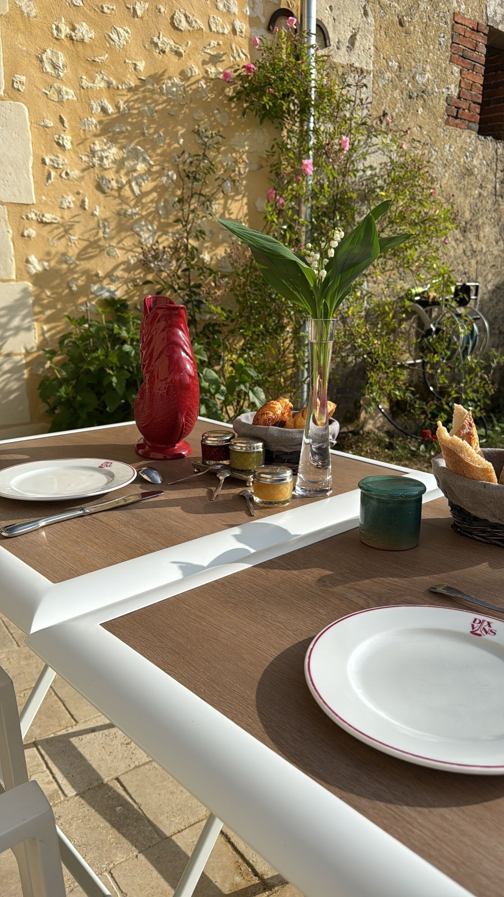 Una mesa al aire libre con platos, cubiertos, un florero con flores y una cesta de pan.