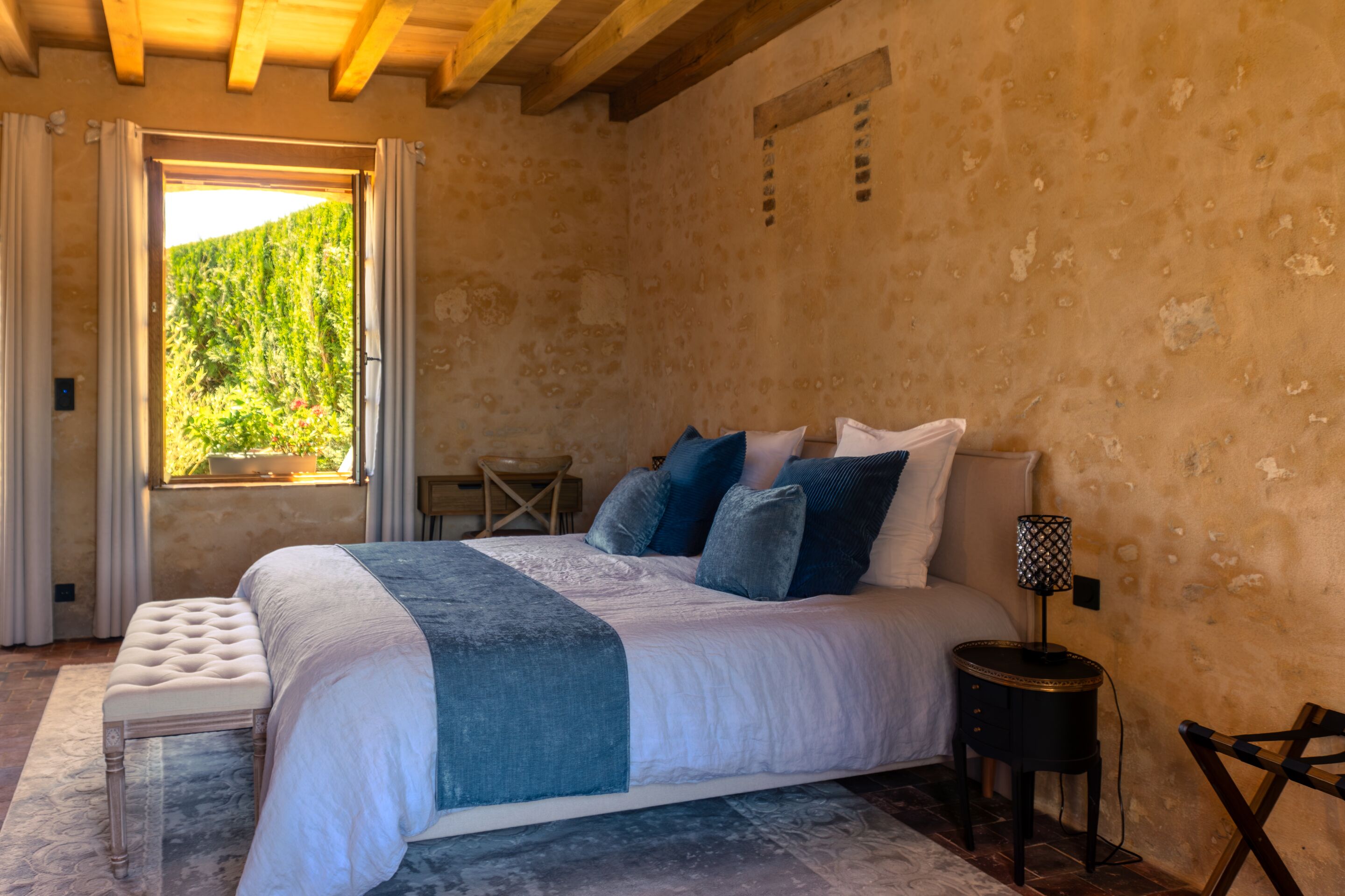 A bedroom with a large bed, a bedside table, a chair, and a window with a view of greenery.