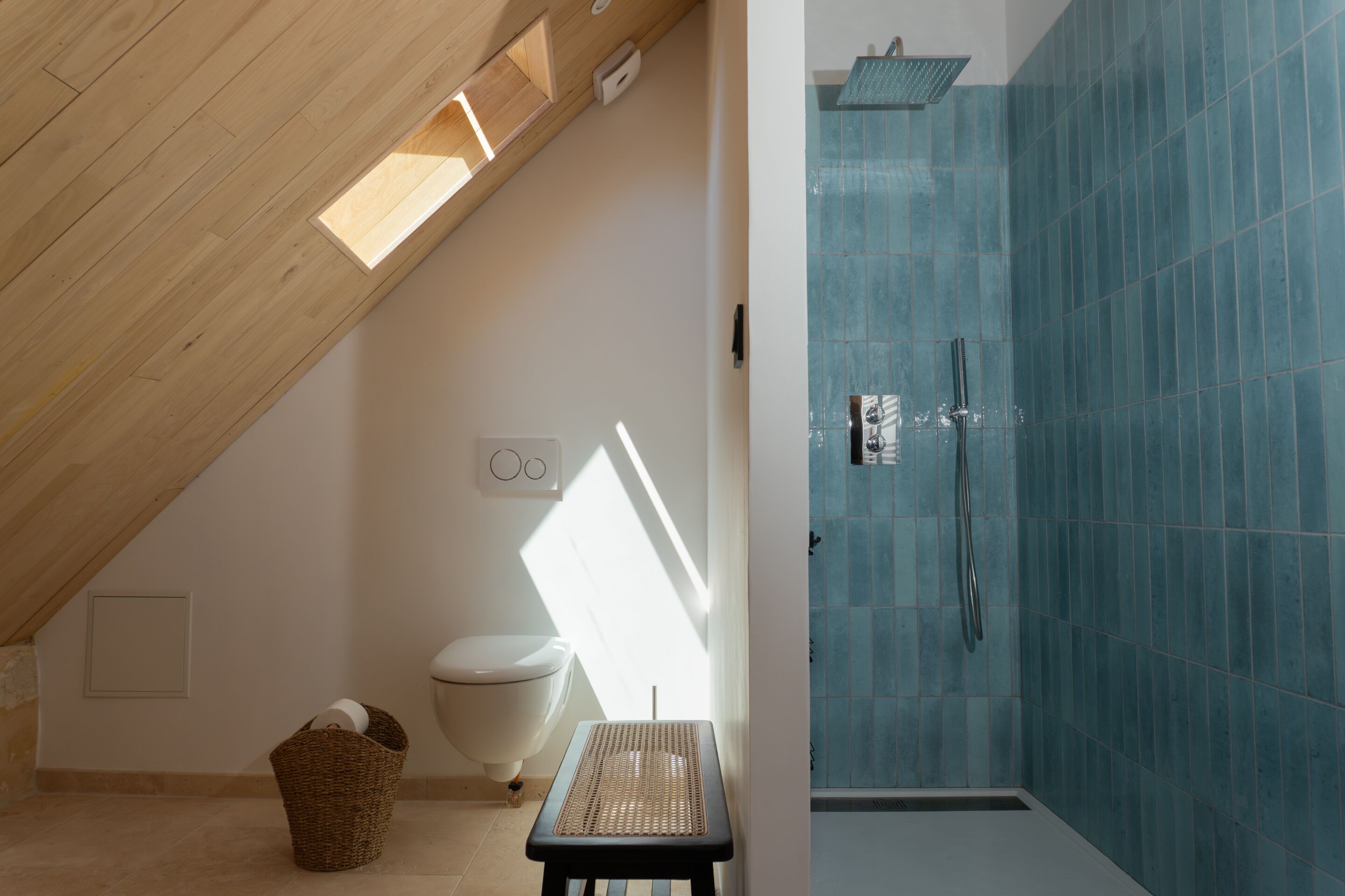 A bathroom with a sloped ceiling, a toilet, a shower with blue tiles, and a woven basket on the floor.