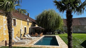 A swimming pool with lounge chairs and a patio area, surrounded by palm trees and a stone building. - RELAI VALDIEU (Courgeon)