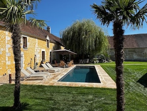 A swimming pool with lounge chairs and a patio area, surrounded by palm trees and a stone building.