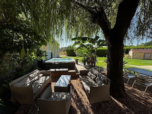 An outdoor seating area with sofas and a hot tub, surrounded by trees and a lawn.